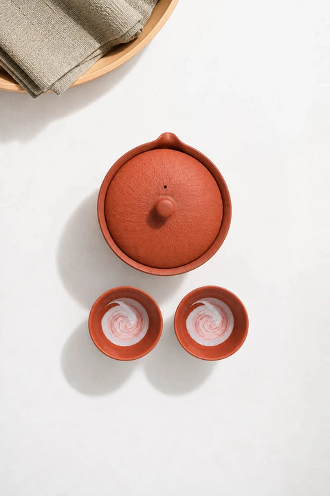 A terracotta clay teapot with its lid on, shown next to two matching small cups with white swirl patterns inside, arranged on a light beige surface with soft shadows.