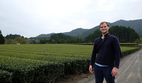 Will Klingner wearing a dark navy quarter-zip pullover and dark pants stands on a dirt path next to a vast green tea plantation, with forested mountains in the background under an overcast sky.