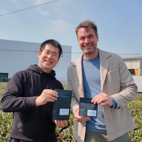 Oishi farmer and Nio Teas' CEO standing outdoors in a green tea plantation holding green tea  pouches with white labels under a blue sky.