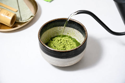 A ceramic matcha tea bowl filled with green matcha powder, with hot water being poured from a black gokuroh kettle. A bamboo whisk and tea towel are visible in the background on a white surface.